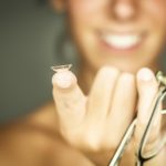 out of focus smiling woman holding up a pair of eyeglasses and a contact lens