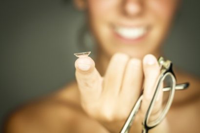 out of focus smiling woman holding up a pair of eyeglasses and a contact lense