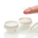 close up of an open contact lens case and a finger holding a contact lens on its tip