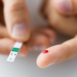 close up of a male finger with a spot of blood and a blood sugar level testing strip