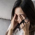 tired woman with glasses rubbing her eyes on sofa